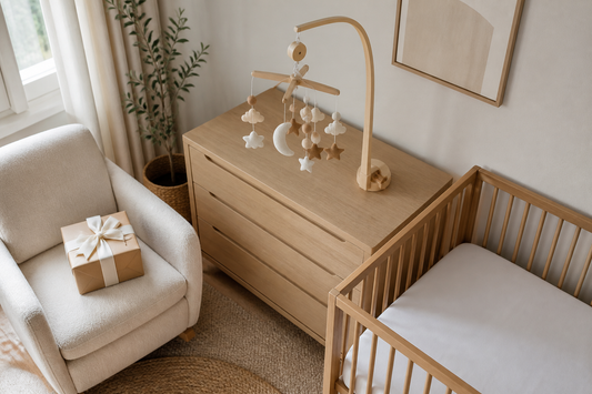 High-angle view of a soft modern UK nursery with a wrapped baby shower gift on a chair, a moon-and-star felt cot mobile on a wooden hanger and a clear light oak cot