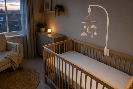 High-angle view of a light oak cot with a secure white wooden mobile hanger and a calm moon-and-cloud cot mobile hanging high above a clear mattress, with a small warm night light on a dresser away from the cot