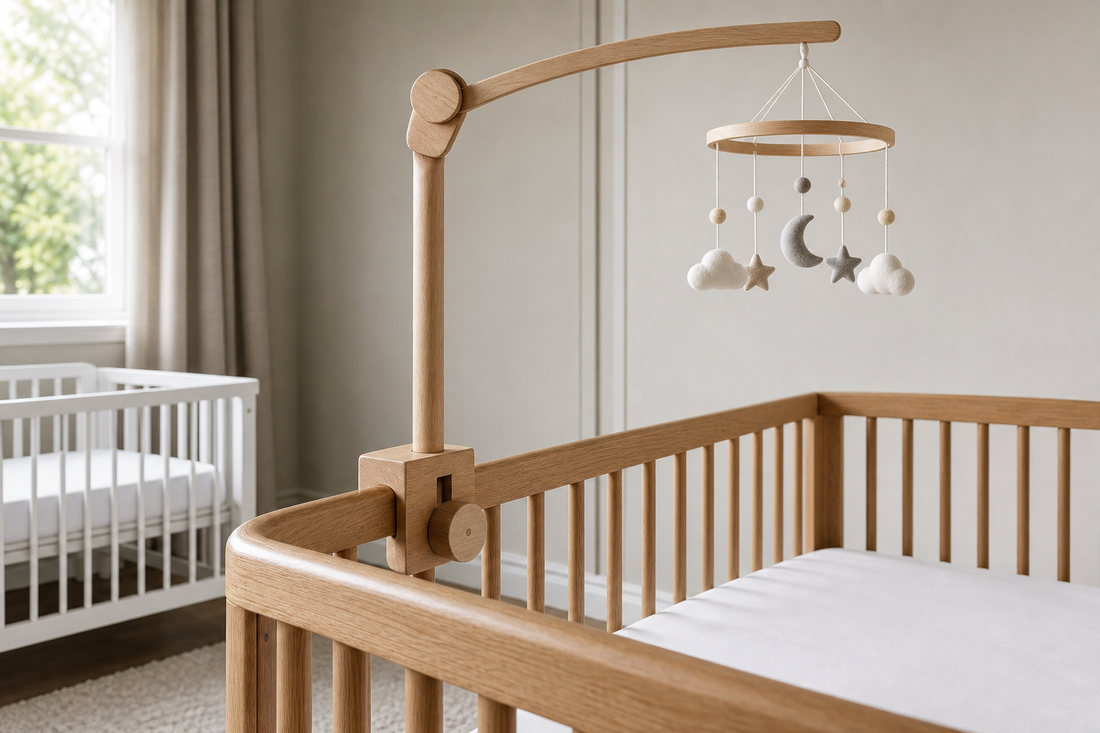 Detail-led cot-side view of a wooden mobile hanger fixed securely to a clear light oak cot rail with a smaller white crib-style sleep space softly visible in the background of a modern UK nursery
