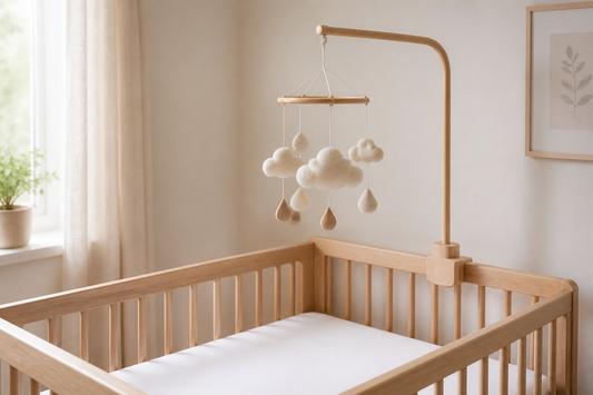 Three-quarter cot-side view of a light oak cot with a wooden mobile hanger holding a simple cloud-and-raindrop cot mobile high above a clear white mattress in a soft modern UK nursery